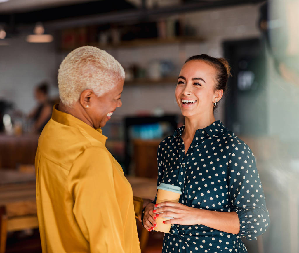An older and younger woman laughing with each other, with young woman holding coffee cup