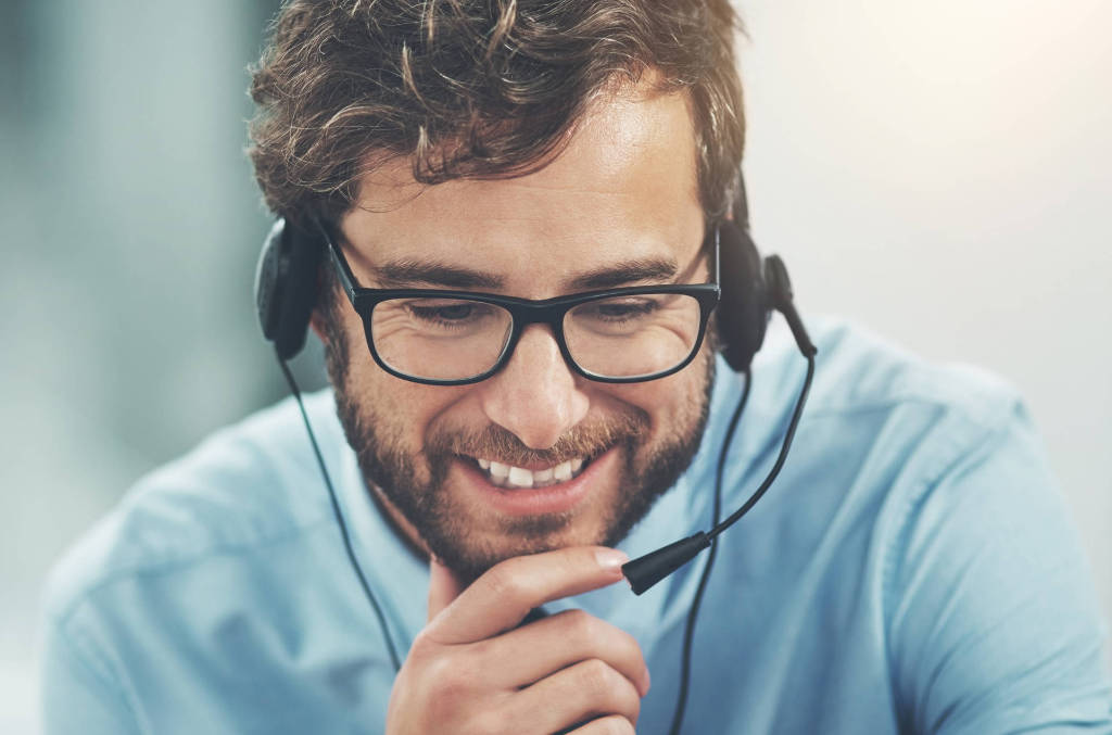 Customer service man with blue shirt and headset leaning forward with chin in hands and smiling