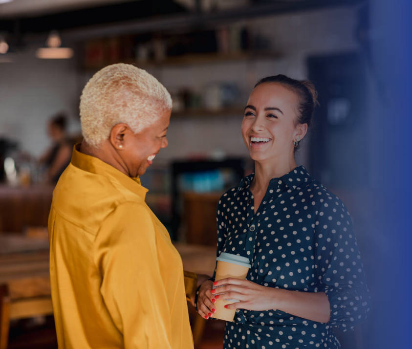 An older and younger woman laughing with each other, with young woman holding coffee cup